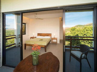 View through sliding glass doors into a bedroom with a wooden bed, nightstand, and a balcony featuring a table with a plant, a chair, and scenic greenery outside.