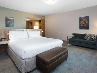 A modern hotel room featuring a large bed with white linens, a gray sofa, and a dark brown ottoman. Artwork hangs on the light gray walls, and a mirrored wall and lamp are visible in the background.