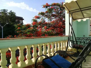Balcony with white railing, black chairs, and a canopy, overlooking vibrant red and green trees against a blue sky.