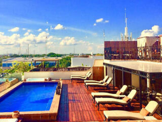 Rooftop pool area with wooden decking, lounge chairs, and a shaded seating area. Cityscape and clear blue sky in the background.