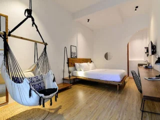 Modern bedroom with hardwood flooring, a large bed, a hammock chair, wall art, a desk with a TV and chair, and minimalist decor.