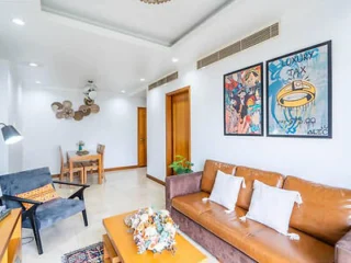 A modern living room with a brown leather sofa, a black armchair, a wooden coffee table, and floral artwork on the walls. A small dining area is visible in the background.