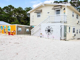 A two-story white building with an external staircase, next to a colorful mural on a yellow wall spelling "PCB." The foreground features a sandy area. Trees are seen in the background.