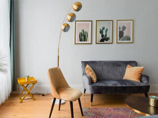 A modern living room features a gray sofa, a beige chair, a yellow side table, a multicolored rug, a tall lamp with three gold shades, and three framed cactus pictures on the wall.