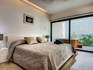 Modern bedroom with a large bed, neutral-toned bedding, two bedside tables with lamps, and a wall-mounted picture. A glass door opens to a patio with a hot tub and greenery outside.