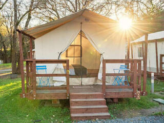 A glamping tent with a wooden deck, two blue folding chairs, and the sun shining through the trees in the background.