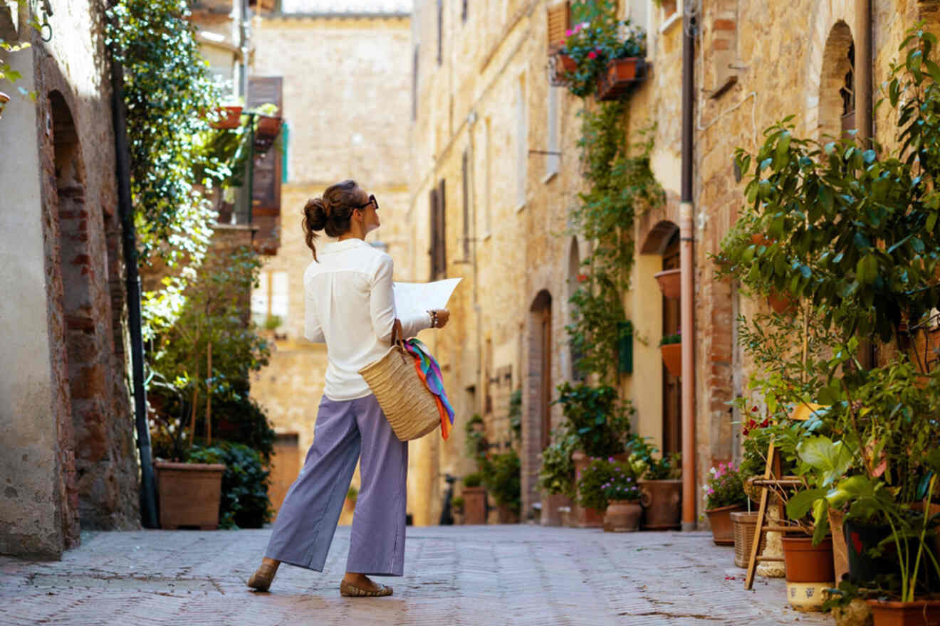 A person stands in a narrow, rustic alleyway lined with plants, holding a map and looking at the surrounding buildings.