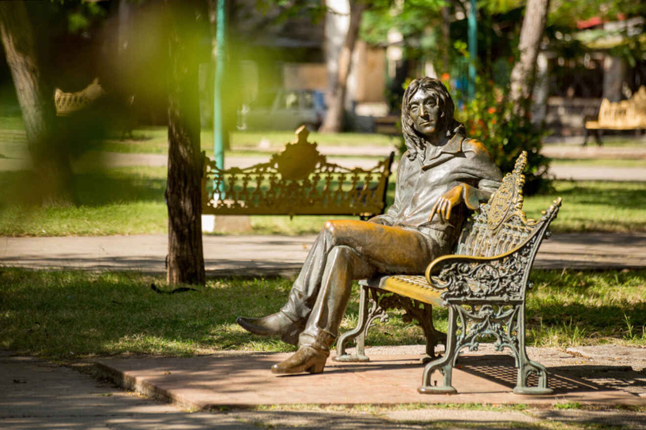 Bronze statue of a seated person on a park bench surrounded by trees and greenery.