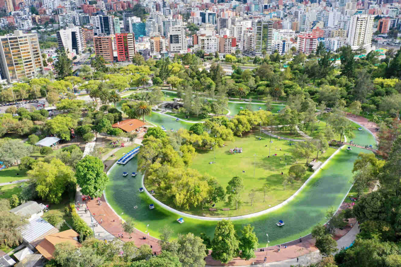 Aerial view of a large, green urban park with winding water features, surrounded by various buildings and trees. People are seen enjoying the open space and walking paths throughout the park.