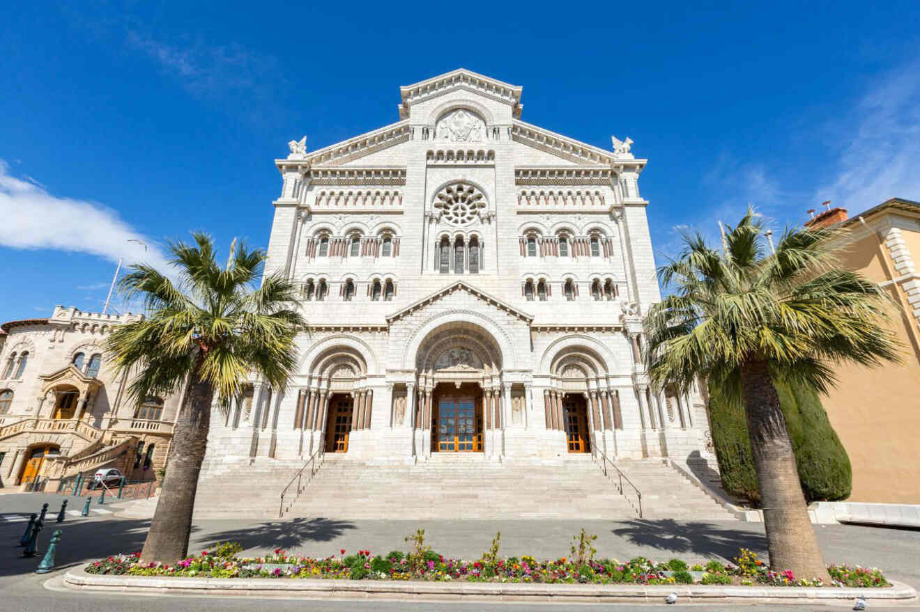 The ornate facade of Saint Nicholas Cathedral in Monaco, framed by palm trees and colorful flowerbeds under a clear blue sky.