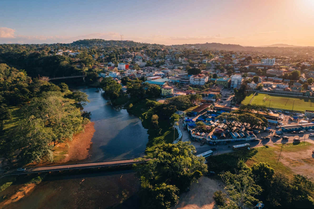 Aerial view of a town at sunset with a river, green areas, residential buildings, and a sports field visible.