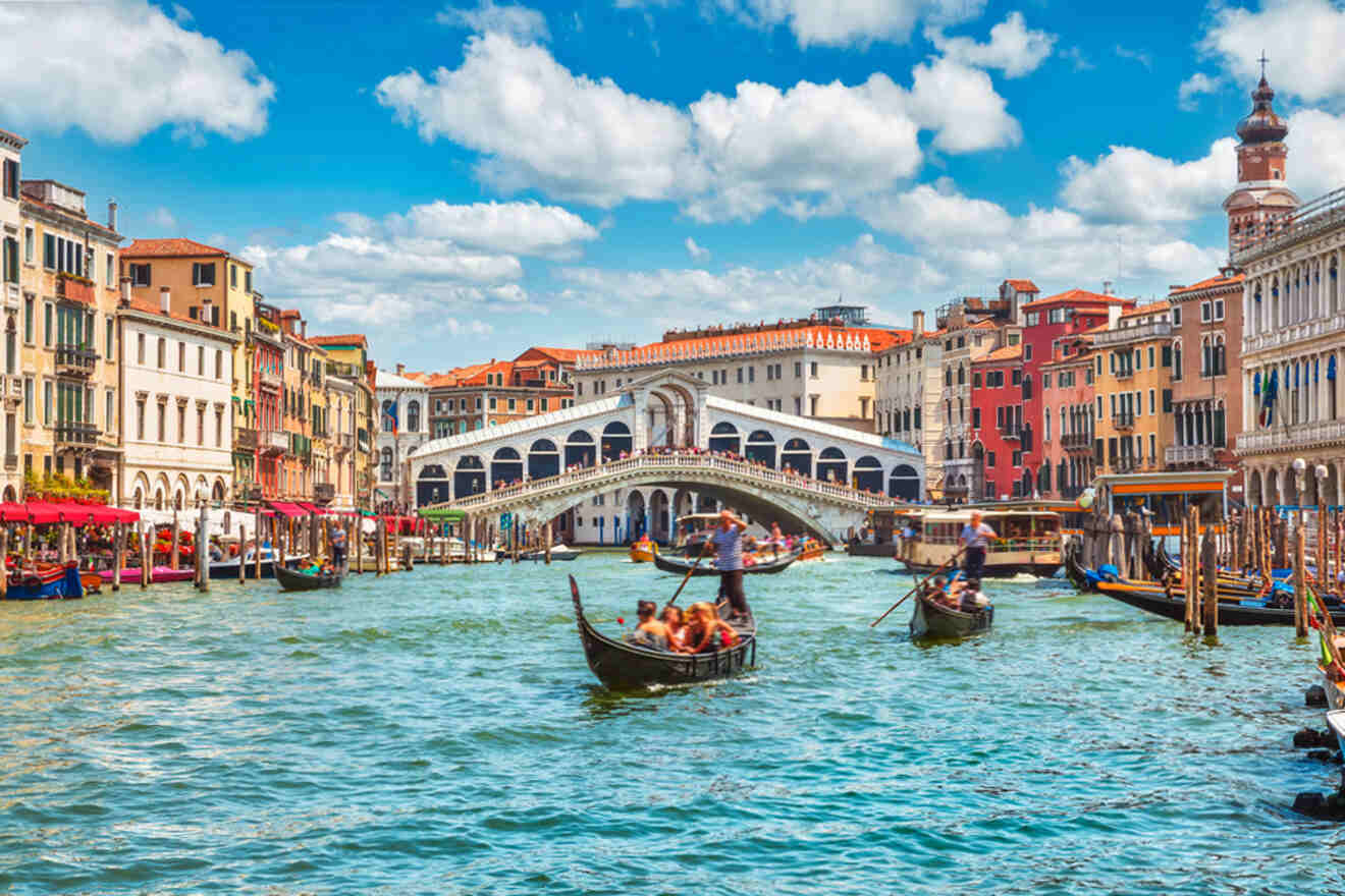 Venetian canal with gondolas in the water, colorful buildings on both sides, and the Rialto Bridge in the background under a partly cloudy sky.