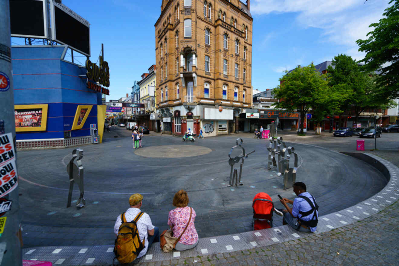 Three people sit on a curb in front of a urban square with metal human figures. A historic brick building and various shops surround the square. Trees and a blue sky are seen in the background.