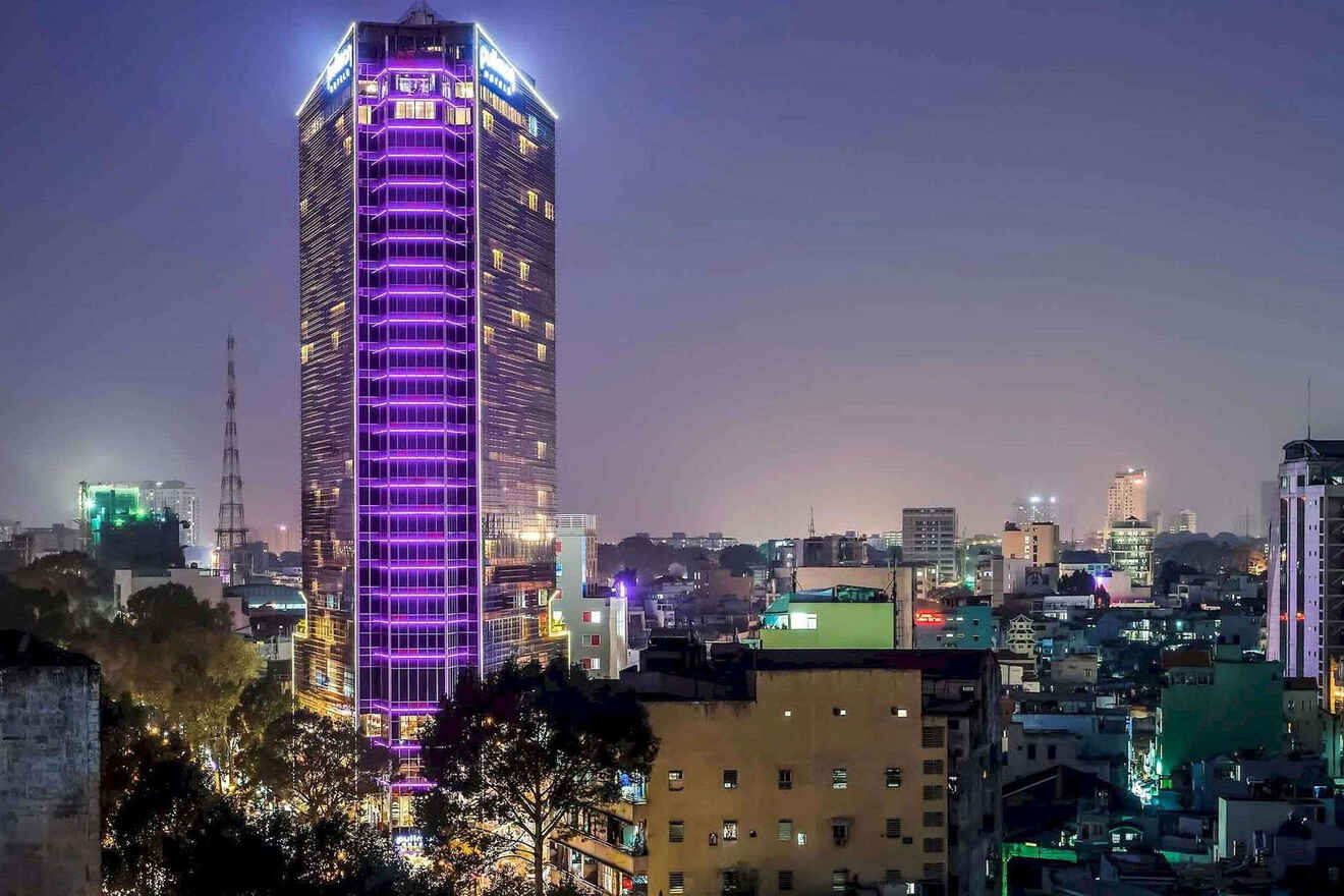 A tall building illuminated with purple lights stands prominently in a cityscape at night, surrounded by lower buildings and distant city lights.