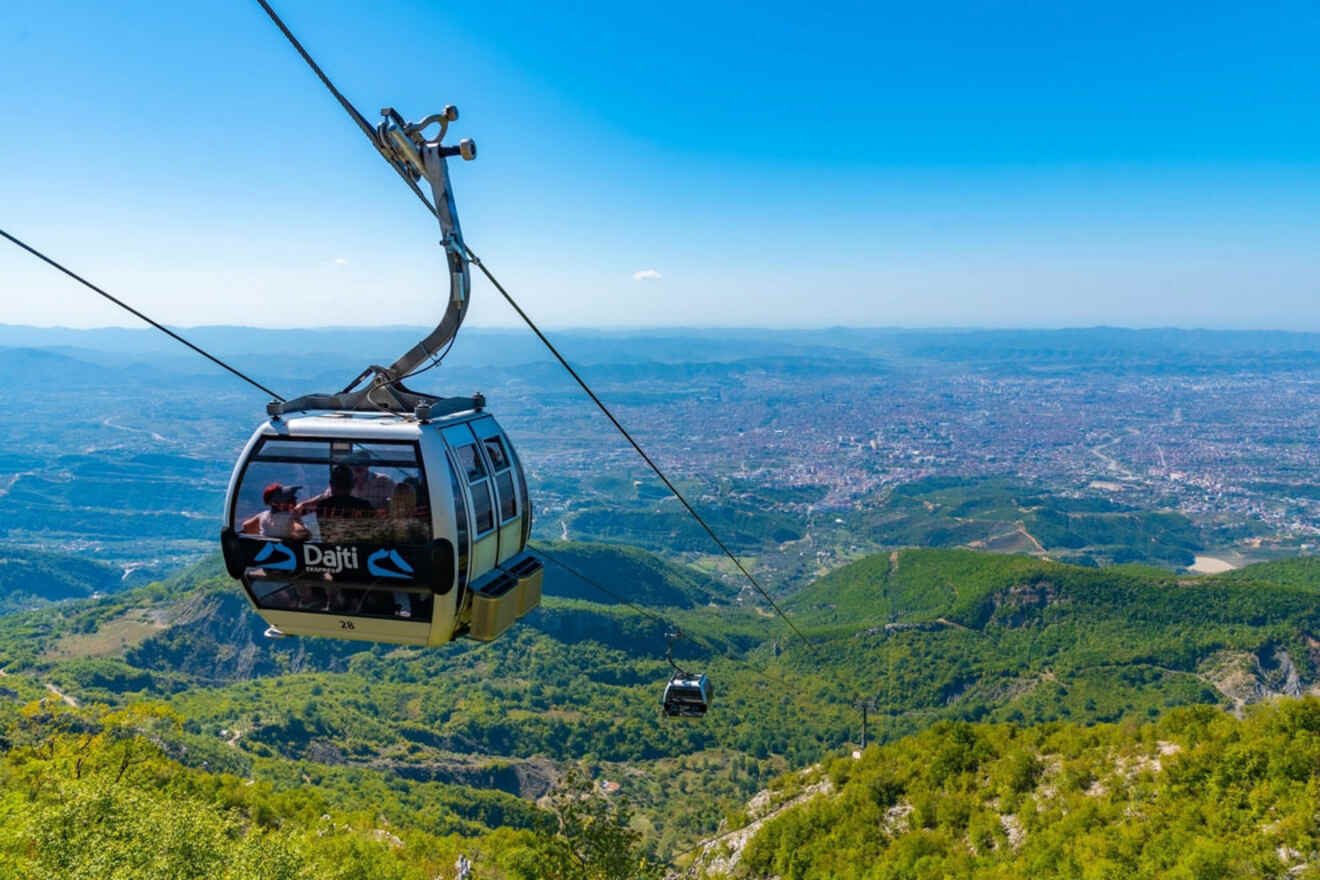 A cable car marked "Dajti" traveling over a lush green landscape with a vast view of Tirana city in the distance.