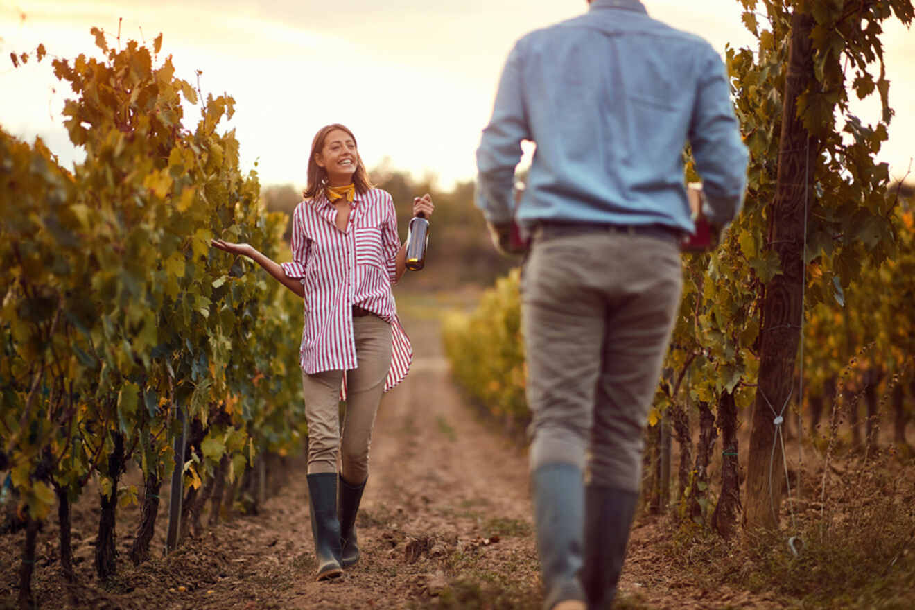 A woman holding a wine bottle and a glass walks towards a man in a vineyard at sunset. Both are dressed in casual clothing and wear boots.