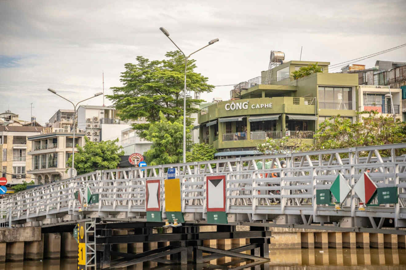 A metal bridge over a river with a green building labeled "Cộng Càphê" in the background, surrounded by other buildings and trees.