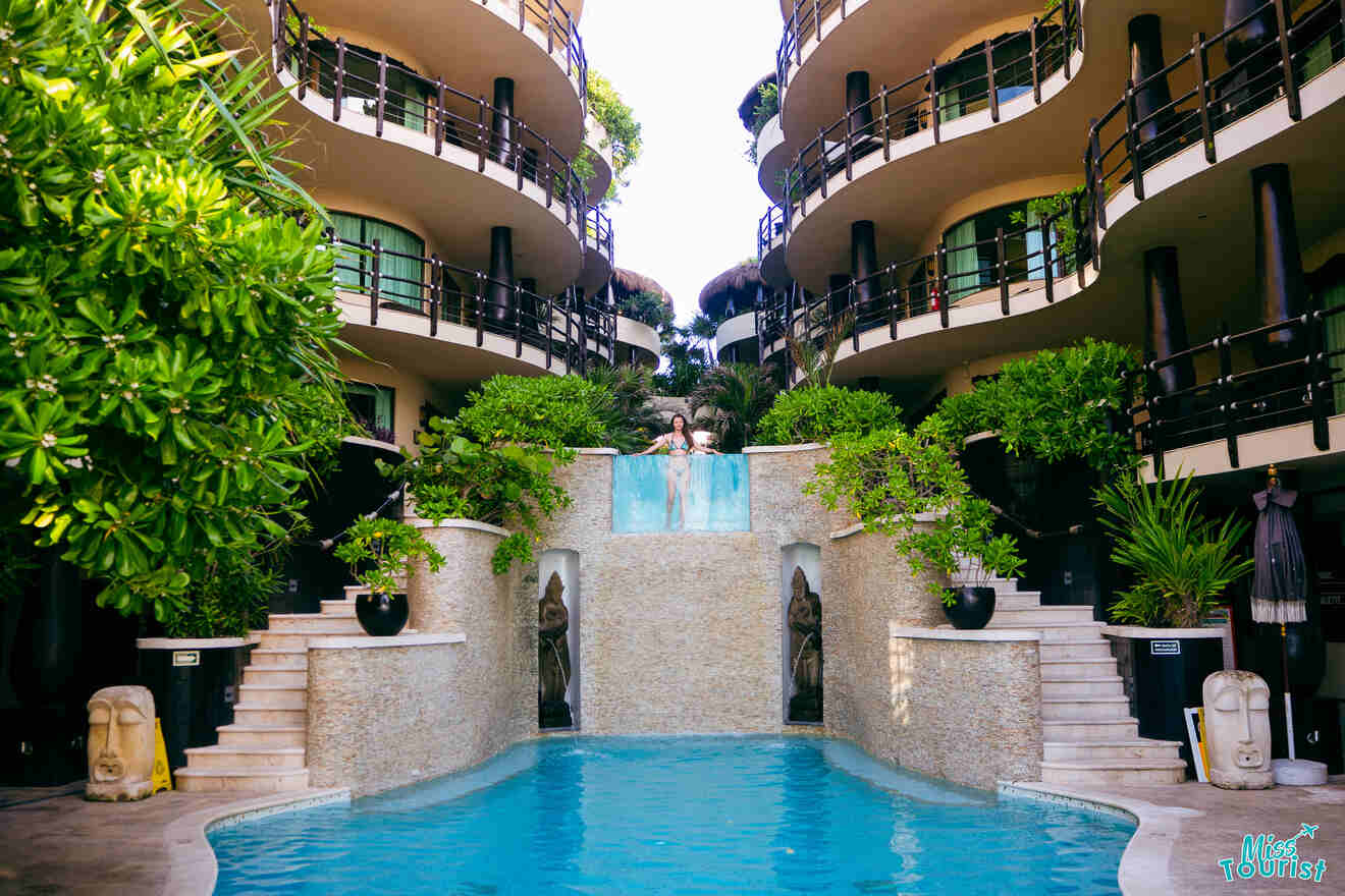 the author of the post in swimming pool with a central waterfall feature is surrounded by modern, multi-level buildings adorned with greenery.