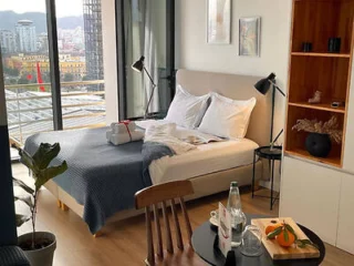 A modern bedroom with a neatly made bed, two reading lamps, a small table set with water and fruit, and a balcony with a city view.
