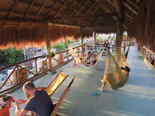 People relax in hammocks and wooden chairs under a thatched roof on a spacious balcony overlooking greenery.