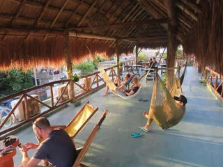 People relax in hammocks and wooden chairs under a thatched roof on a spacious balcony overlooking greenery.