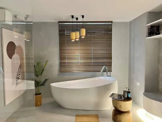 Modern bathroom features a white freestanding bathtub, wooden blinds, indoor plant, framed artwork, and a side table with toiletries against light gray walls.