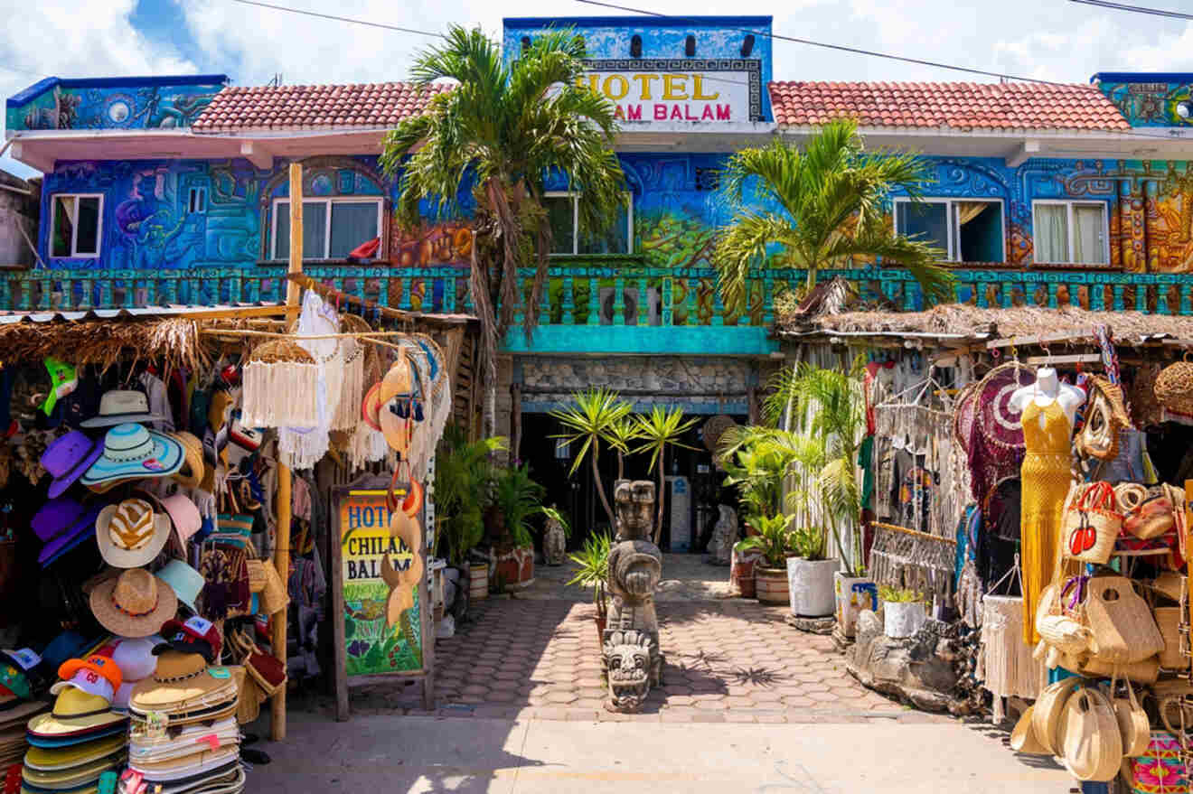 A colorful hotel entrance flanked by two market stalls selling hats, bags, and souvenirs. Two palm trees are in front of the hotel building, which is painted with vibrant murals.