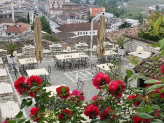 Outdoor terrace with empty tables and chairs, surrounded by red flowers, overlooks a cityscape with various buildings and greenery in the background.