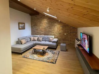 A cozy living room features a grey sectional sofa with tan pillows, a decorative rug, a coffee table, and a wall-mounted TV. The space has a slanted wooden ceiling and a brick accent wall.
