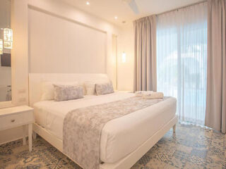 A modern bedroom featuring a large bed with white and gray bedding, a nightstand, and large windows with sheer curtains. The room is well-lit and has patterned floor tiles.