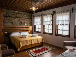 A cozy bedroom with a double bed, wooden headboard, and three windows with lace curtains. Two towels are neatly placed on the bed. The room features a decorative rug and a stone accent wall.