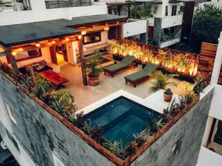 A rooftop patio featuring a small pool, two lounge chairs, a seating area, and various plants along the edges. The space is illuminated with ambient lighting.