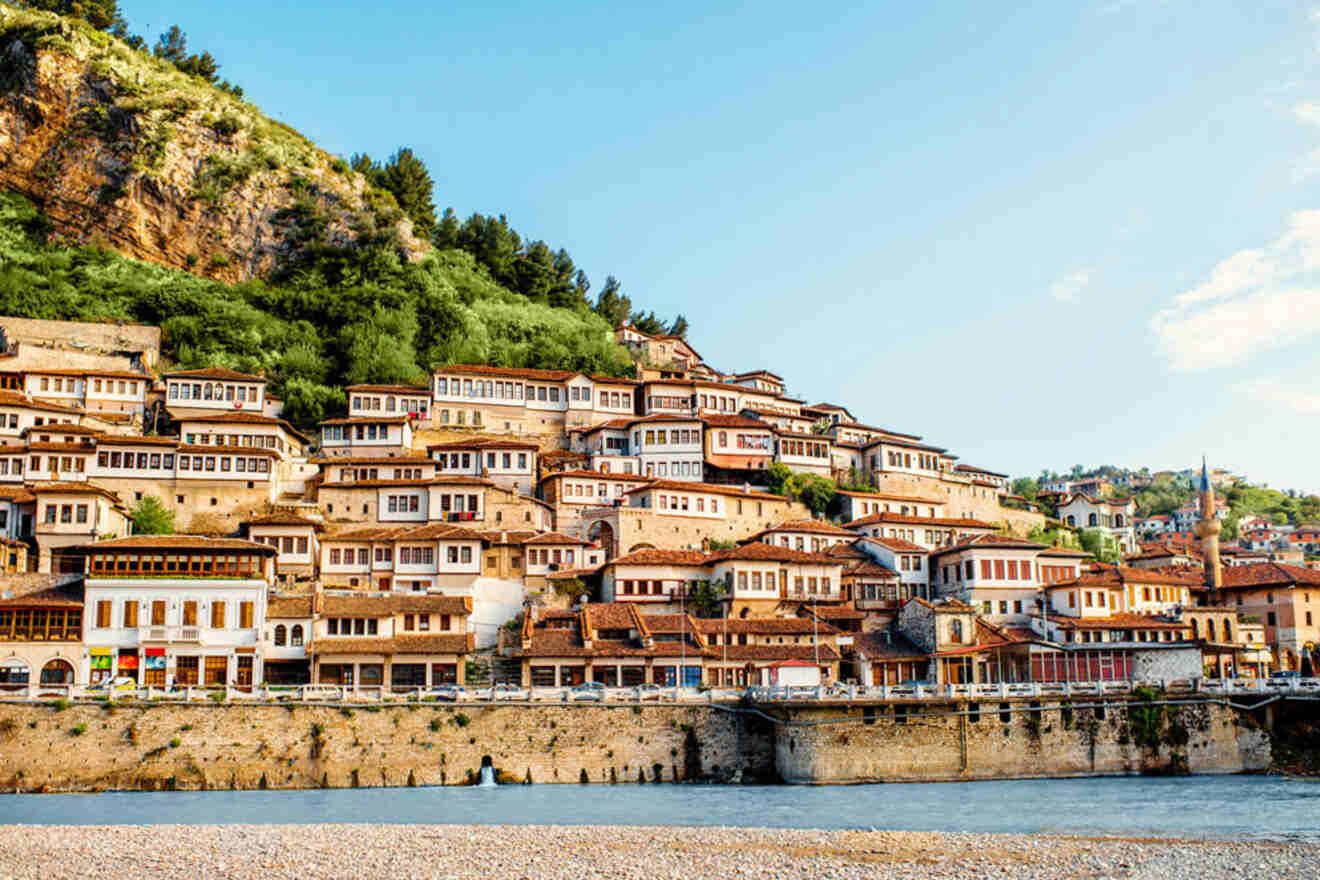 View of a hillside town featuring numerous traditional houses with red rooftops and white walls. A stone bridge spans across the river in the foreground. Greenery covers the hilltop above the town.