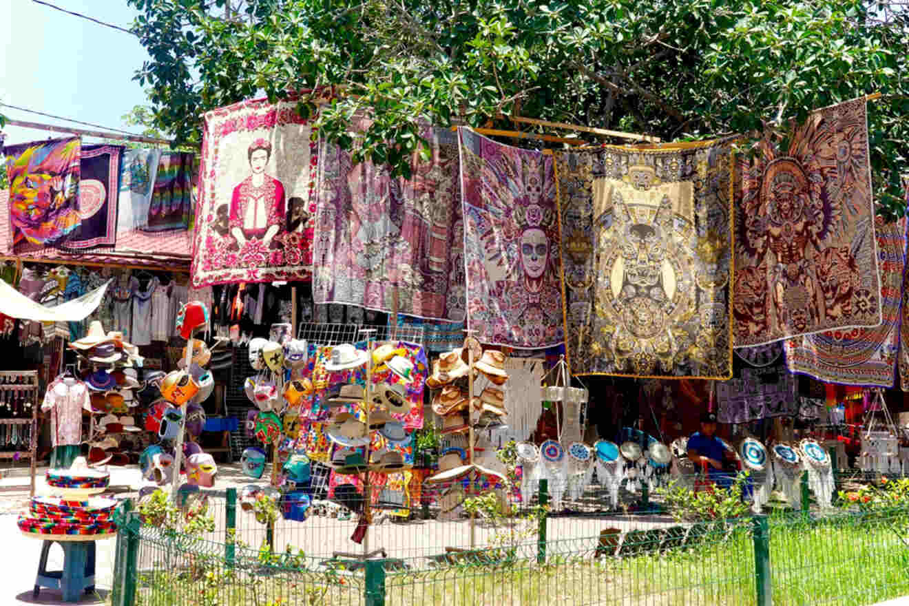 Outdoor market stall displaying colorful tapestries, hats, and various crafts hung up under tree branches. The background shows additional stalls with assorted merchandise.