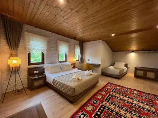 A cozy bedroom with wooden ceiling, two double beds, decorative rug, and windows with sheer curtains. Lamps on bedside tables and a desk with a chair are also present.