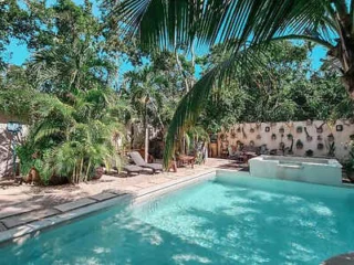 A serene outdoor pool area surrounded by lush greenery and tall palm trees, featuring poolside lounge chairs and a wooden fence adorned with potted plants in the background.