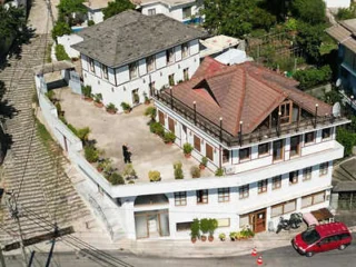 Aerial view of a residential property with two multi-story houses, a courtyard, potted plants, and a person standing in the courtyard. A staircase and road are visible on the left side.