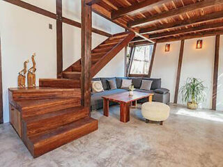 A modern living room with a wooden staircase, grey sectional sofa, wooden coffee table, potted plant, round ottoman, and bird sculptures on the stairs. The floor is concrete and walls are white.