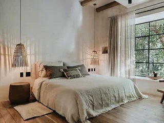 A cozy bedroom features a bed with beige linens and decorative pillows, flanked by two pendant lights. A large window with curtains offers a view of trees, and wooden furniture complements the room.