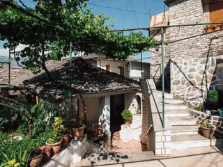 Stone house courtyard with potted plants and greenery, partially covered by a trellis. Stone stairs lead to an upper level. The sky is clear and blue.