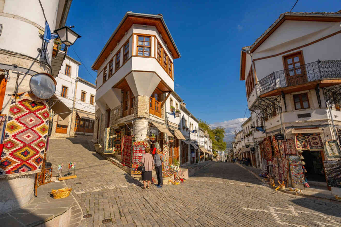 A cobblestone street is flanked by traditional white buildings with wooden accents. Colorful rugs and crafts are displayed outside shops. Two people stand near a shop. Bright, sunny day.