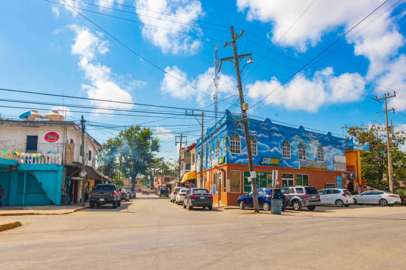 Street view of a small town with colorful buildings, parked cars, and power lines overhead under a blue sky with scattered clouds.