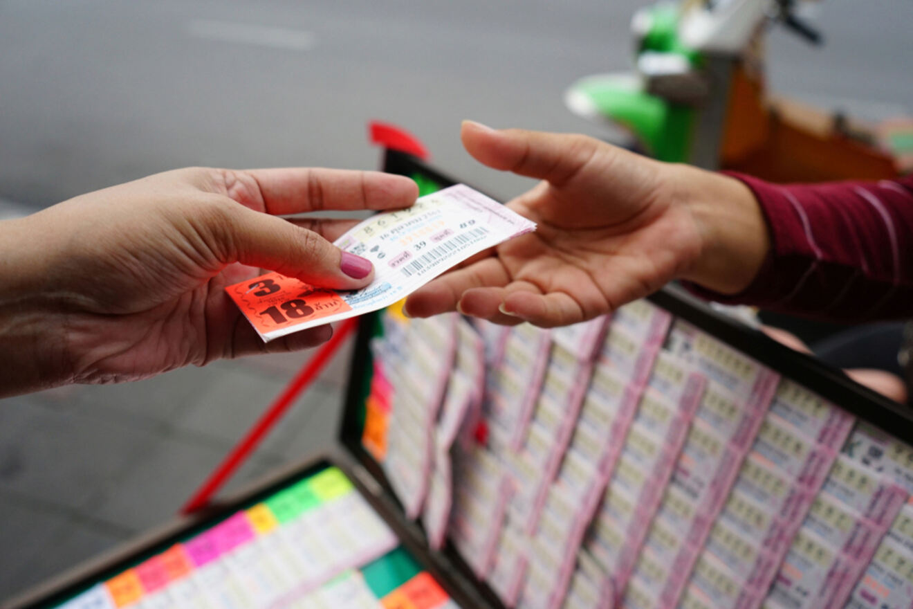 A close-up of hands exchanging money and a lottery ticket, with a display of colorful lottery tickets visible in the background.