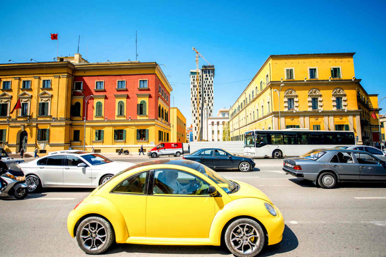 A vibrant yellow car drives through a bustling street in Tirana, surrounded by historic buildings and traffic.