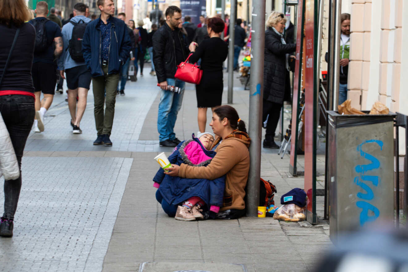 A woman sitting on the street with a child in her lap, holding a book, surrounded by pedestrians walking by in a busy urban area.