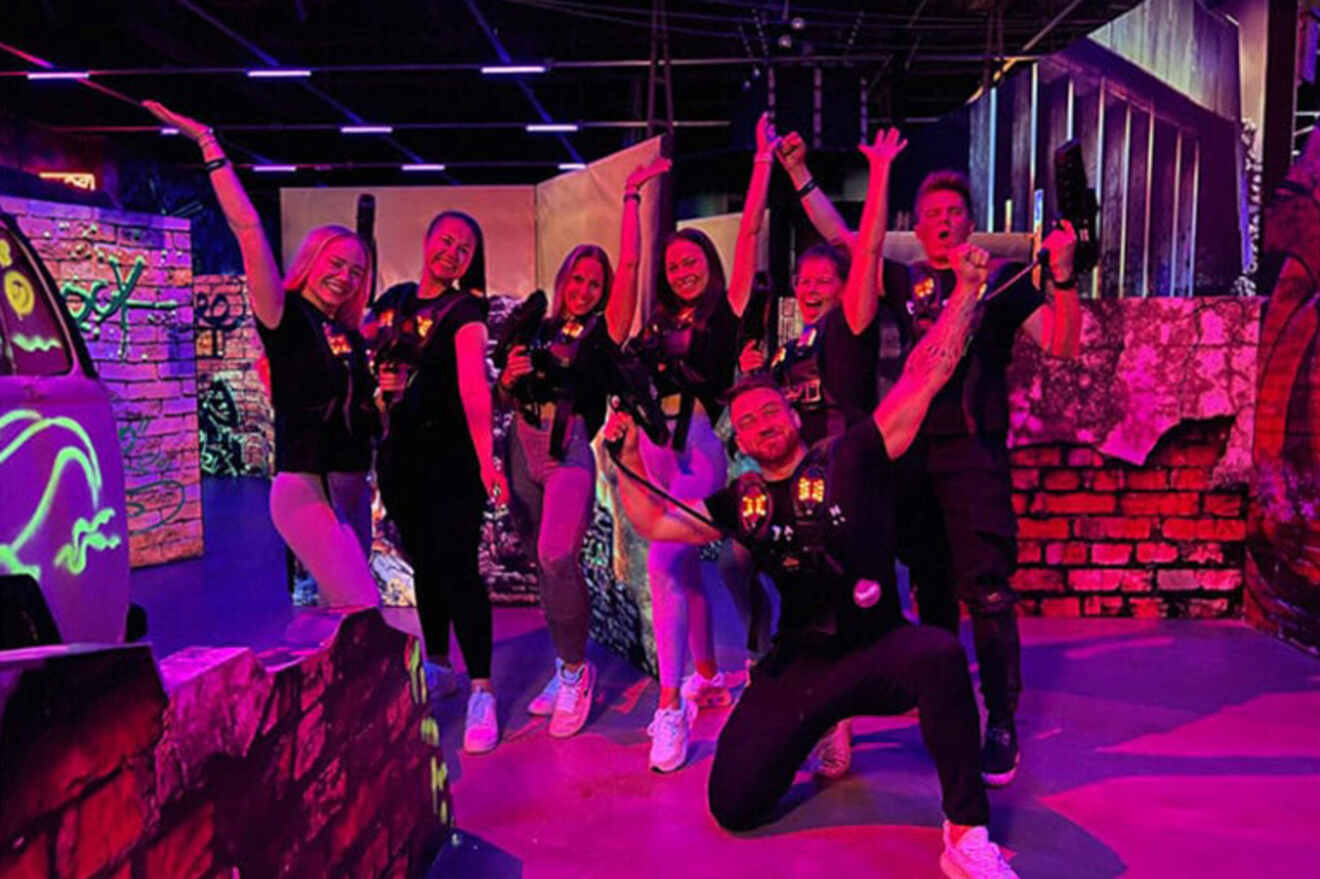 A group of people in a laser tag arena wearing vests and holding laser guns pose enthusiastically for a photo. The setting features neon lights and vivid decorations.