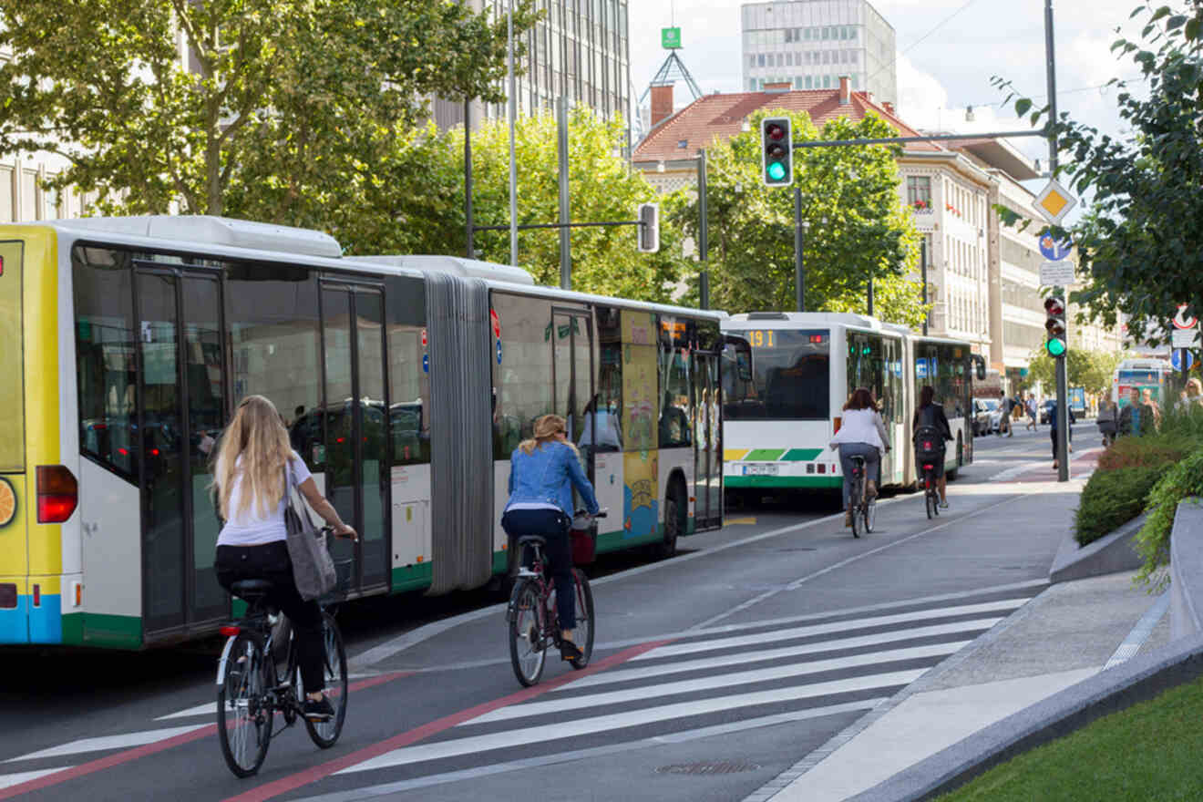Two buses stopped at a traffic light as three cyclists ride in a dedicated bike lane beside them on a city street surrounded by buildings and trees.