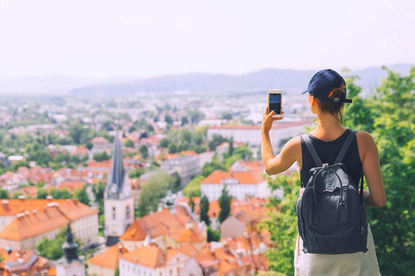 A person with a backpack and cap takes a photo of a cityscape featuring buildings with red rooftops and a church steeple.