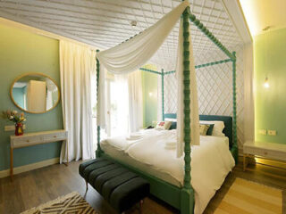 A well-lit bedroom with a teal canopy bed, white drapes, and pillows. The room has mint green walls, a circular mirror, side tables, a black bench at the foot of the bed, and wooden flooring.