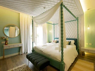 A well-lit bedroom with a teal canopy bed, white drapes, and pillows. The room has mint green walls, a circular mirror, side tables, a black bench at the foot of the bed, and wooden flooring.